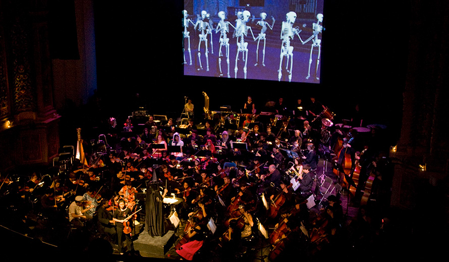 Orchestra on stage beneath a giant screen projecting dancing