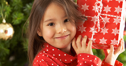 Smiling pajama-clad little girl holding a wrapped gift in front of a Christmas tree